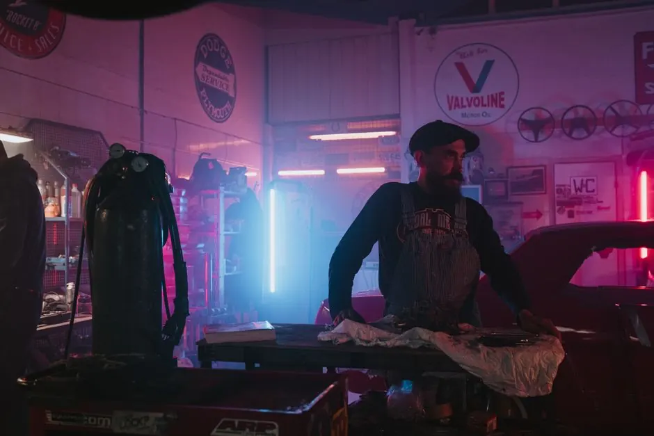 Mechanic works in a vintage garage with neon lighting and classic cars.