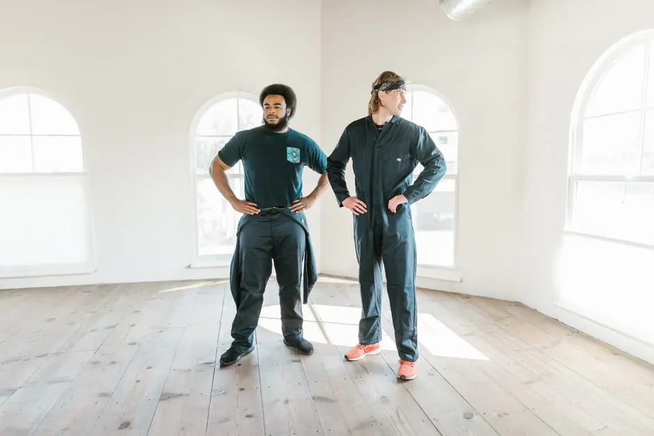 Two men standing confidently in a bright room with hands on waist, wearing dark jumpsuits.